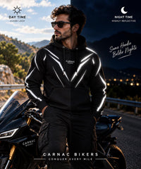 A man wearing a black hooded jacket with reflective white geometric stripe details stands beside a motorcycle on a road at dusk. The image includes text overlays reading 'DAY TIME CLEAN LOOK', 'NIGHT TIME HIGHLY REFLECTIVE', 'Same Hoodie Bolder Nights', and 'CARNAC BIKERS CONQUER EVERY MILE'.