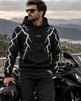 A man wearing a black hoodie with a white lightning-bolt pattern and the brand name 'CARNAC RIDE' on the chest, standing next to a black motorcycle with a scenic mountain background.