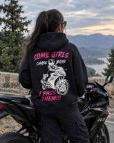 A person standing on a black motorcycle, photographed from the back, wearing a black hoodie with a large graphic print on the back. The print features pink and white text reading 'SOME GIRLS CHASE BOYS I PASS THEM!!!' along with a white motorcycle rider illustration.