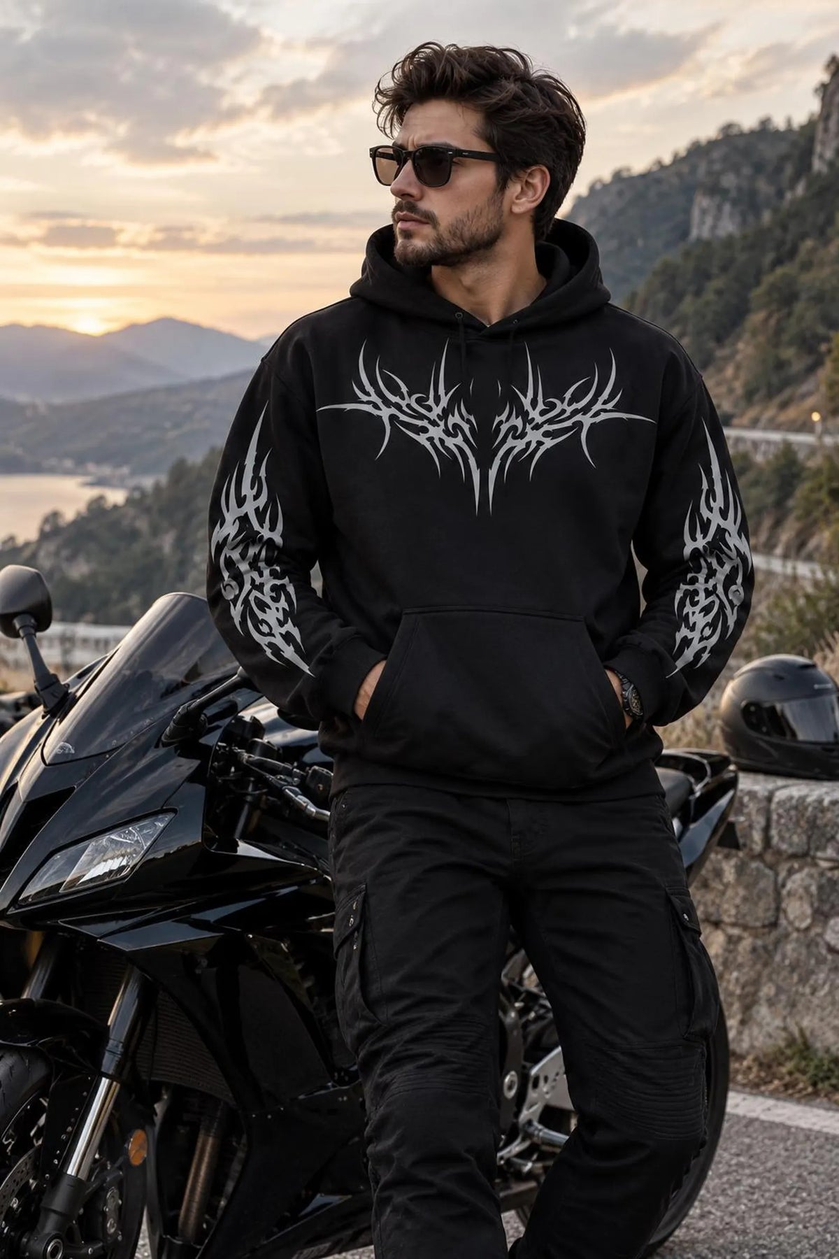 A man wearing a black hoodie with a large white tribal graphic on the chest and sleeve prints, standing next to a black motorcycle in an outdoor mountain setting at sunset.