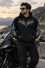 A man wearing a black hoodie with a large white tribal graphic on the chest and sleeve prints, standing next to a black motorcycle in an outdoor mountain setting at sunset.