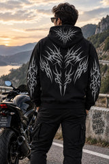 A man stands with his back to the camera wearing a black hoodie with a large white tribal wolf graphic on the back and sleeves, standing next to a black motorcycle on a road with a scenic mountain and lake background at sunset.