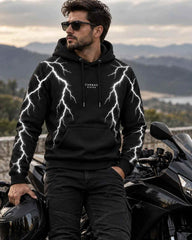 A man wearing a black hoodie with a white lightning-bolt pattern and the brand name 'CARNAC RIDE' on the chest, standing next to a black motorcycle with a scenic mountain background.