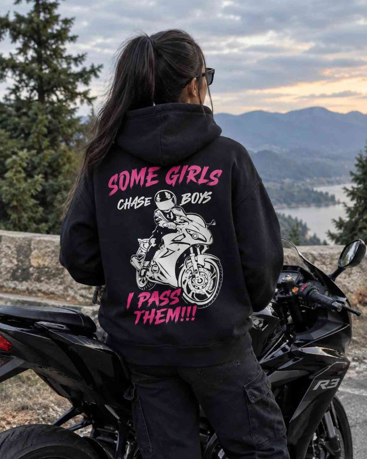 A person standing on a black motorcycle, photographed from the back, wearing a black hoodie with a large graphic print on the back. The print features pink and white text reading 'SOME GIRLS CHASE BOYS I PASS THEM!!!' along with a white motorcycle rider illustration.